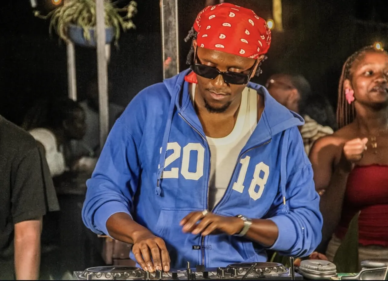 DJ in a red paisley bandana, dark sunglasses and bright blue hoodie mixing at night with the crowd dancing behind
