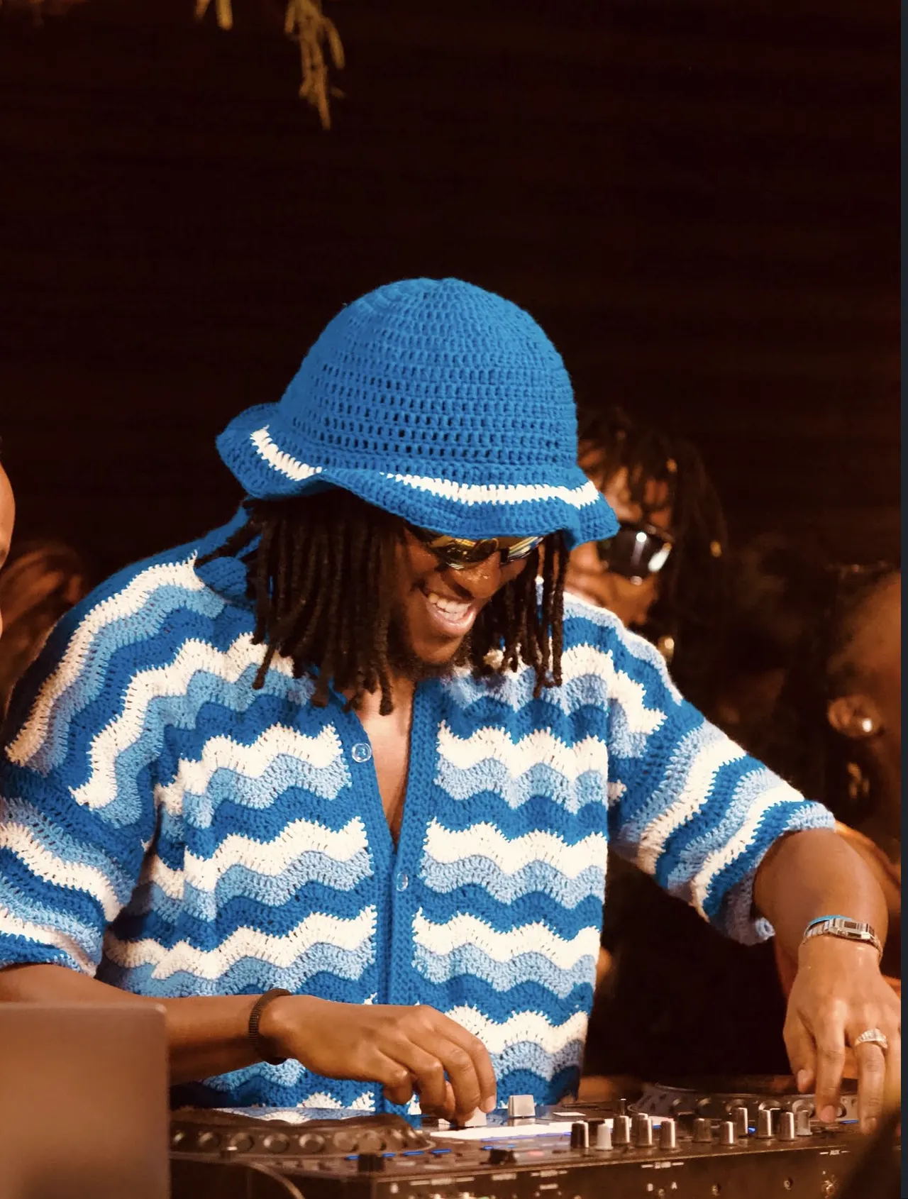 DJ grinning at the decks in a blue-and-white crocheted wave-pattern bucket hat and cardigan, under warm amber light