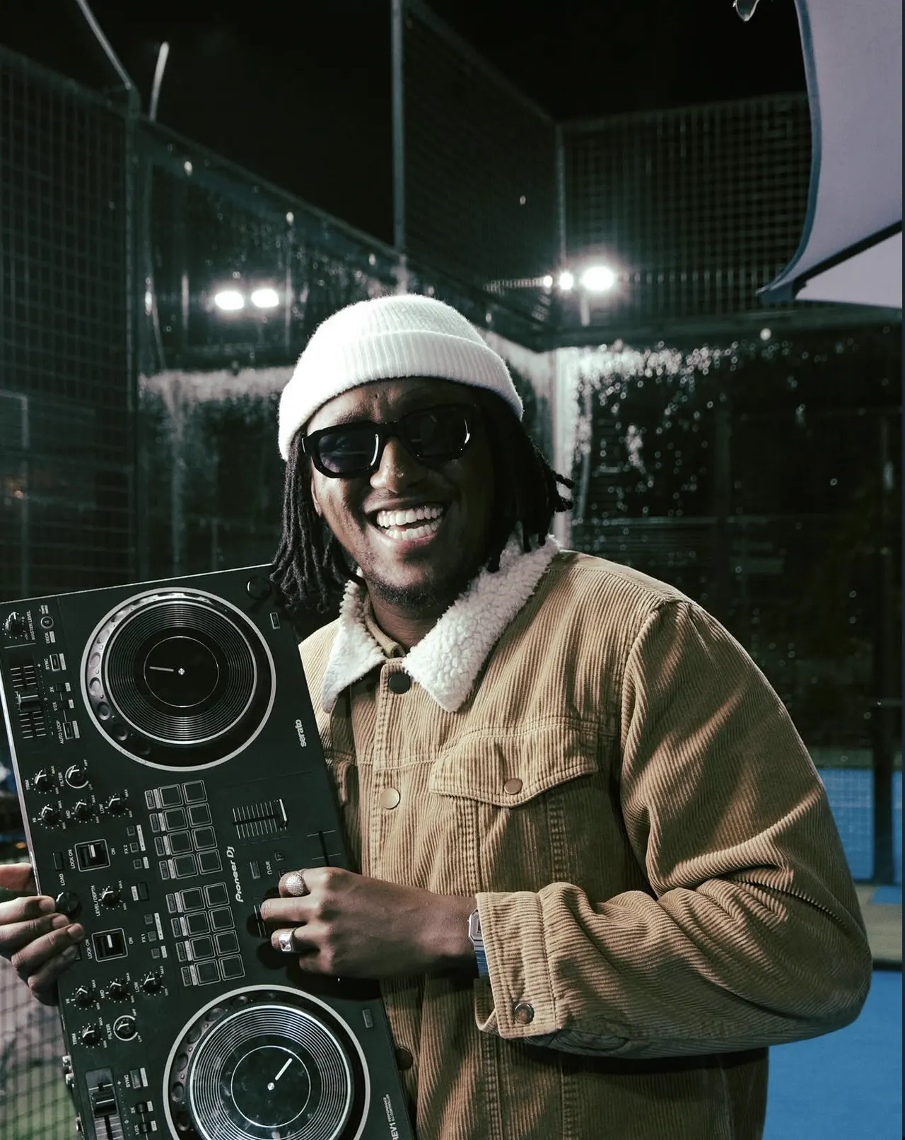 DJ in a white beanie and tan corduroy jacket smiling big while holding a Pioneer DJ controller under floodlights